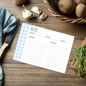 Puede incluir: Una tarjeta de recetas con un borde de cuadros vichy azul claro. La tarjeta tiene espacios para el tiempo de preparación, los ingredientes, el tiempo de cocción, las porciones y las instrucciones. Sobre una superficie de madera con un cuchillo, ajo, patatas y hierbas.