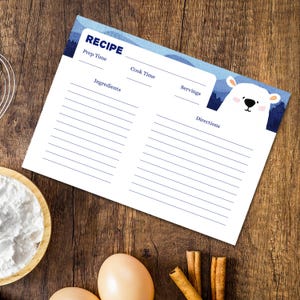 May include: A recipe card with a blue and white mountain design and a cute polar bear illustration. The card includes sections for prep time, cook time, ingredients, servings, and directions. The card is on a wooden surface with baking ingredients.