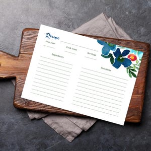 May include: A white recipe card with the word "Recipe" in elegant script. The card features a watercolor floral design in shades of blue, green, and red. The card is on a wooden cutting board with a gray cloth.