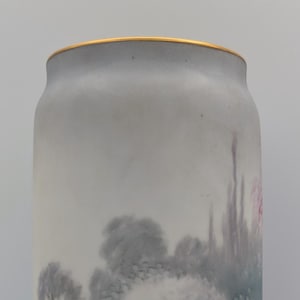 May include: A tall, cylindrical vase with a painted landscape scene. The vase has a light grey background and a gold rim. The landscape depicts a brick wall with hanging vines and pink flowers.