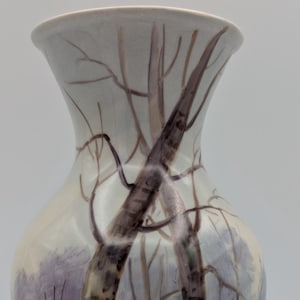 May include: A tall, off-white ceramic vase with a winter landscape scene. The vase has a light blue base and rim, with a painted design of trees, snow, and a muted colour palette. The artwork depicts a serene winter scene.