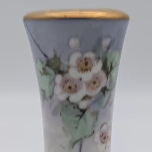 May include: A small, hand-painted porcelain vase with a gold rim. The vase features a delicate floral design with white and pink flowers, green leaves, and a light blue background.