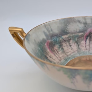 May include: A porcelain bowl with a painted landscape scene. The bowl has a light pink exterior and gold-coloured handles. The interior features a painted scene with pink flowers, trees, and a body of water.