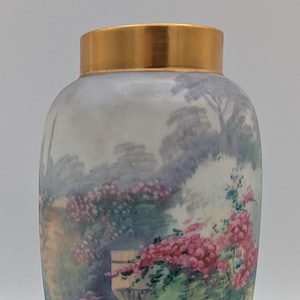 May include: A decorative vase with a gold rim and a painted garden scene. The scene features pink roses, a stone wall, and a bench. The vase has a rounded shape and a blue base.