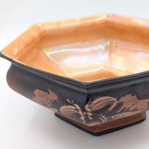 May include: An octagonal bowl with a black exterior and a peach-coloured interior. The bowl has a floral design in a bronze colour on the exterior. The bowl is a decorative item.