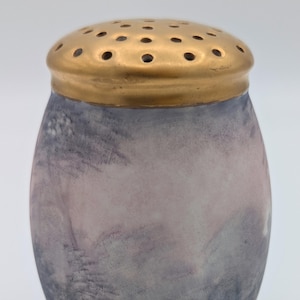 May include: An oval salt shaker with a gold-coloured, perforated lid. The body of the shaker has a watercolour-like design in shades of lavender, grey, and cream. The design gives the shaker a soft, artistic appearance.