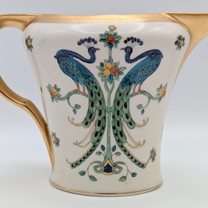 May include: Cream ceramic jug with a gold handle and rim. The jug features a design of two blue peacocks perched on a branch with green and yellow accents. A decorative piece for serving.