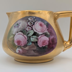 May include: A gold-coloured jug with a floral design. The jug features a painted oval with pink roses, green leaves, and white flowers. The handle is square and the jug has a gold rim.