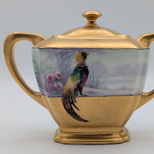 May include: A gold-coloured sugar bowl with a lid and two handles. The bowl features a painted scene of a colourful bird and pink flowers against a landscape background. The lower portion of the bowl has a textured design.