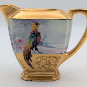 May include: A vintage creamer with a gold rim and handle. The body features a colorful pheasant illustration against a winter landscape. The base has a gold floral pattern. The creamer is a decorative piece.