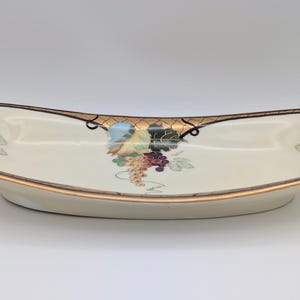 May include: Cream-coloured, oval serving dish with a gold rim and grape cluster design. The dish has a curved shape, likely ceramic or porcelain. The dish is approximately 30 cm long.