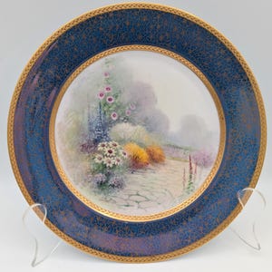 May include: A decorative plate with a blue border and gold floral detailing. The centre of the plate features a garden scene with flowers and a stone path. The plate is displayed on a clear stand.