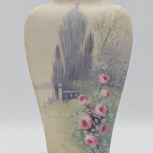May include: A cream-coloured vase with a hand-painted landscape scene. The vase features a design of pink roses and a landscape with trees and a fence. The top and bottom rims have a gold trim.