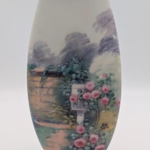 May include: An oval vase with a painted garden scene. The vase has a light blue base and a gold rim. The garden scene depicts pink roses, a stone wall, and trees in shades of green and purple.