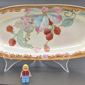 May include: Oval serving dish with a gold rim and handles. The dish has a hand-painted design of raspberries, pink flowers, and green leaves. The dish is supported by a metal stand. A small Lego figure is in the foreground.