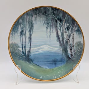May include: A decorative plate with a landscape scene depicting a lake, trees, and mountains. The plate has a gold rim and is displayed on a clear stand. The artwork is predominantly in shades of blue and green.