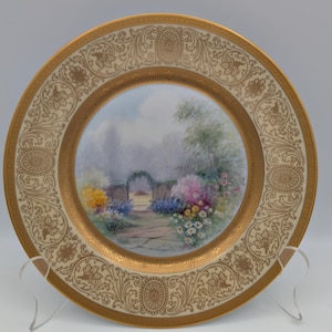 May include: A decorative plate featuring a hand-painted garden scene. The central image shows a path leading to an arched gate, surrounded by flowers. The rim has a gold filigree design on a cream background. The plate is displayed on a clear stand.