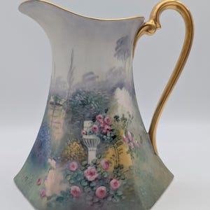 May include: A porcelain jug with a gold handle and rim, decorated with a hand-painted garden scene. The design includes pink roses, a white pillar, and a soft colour palette of greens, blues, and yellows. The jug has a distinctive, faceted shape.