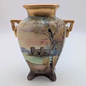 May include: A small, decorative vase with a landscape painting. The vase has a gold rim and handles, and a base with three feet. The painting depicts a castle, trees, and a colorful sky. The vase is likely made of ceramic.
