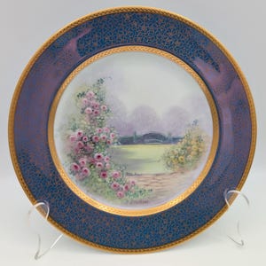 May include: A decorative plate with a blue border featuring a gold floral pattern. The centre of the plate displays a hand-painted garden scene with pink roses, a bridge, and a pathway. The plate has a gold rim.