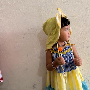 May include: A child wearing a textured lime green sun hat with ties and a tiered dress featuring a blue and white gingham bodice, a yellow and white striped middle section, and a light blue and white gingham hem with yellow trim, holding a lollipop.