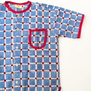 May include: A short-sleeved button-down shirt featuring a light blue grid pattern on a cream background with small red squares at each intersection. The shirt has a red collar, red sleeve cuffs, a red hem, and a single chest pocket with red trim.