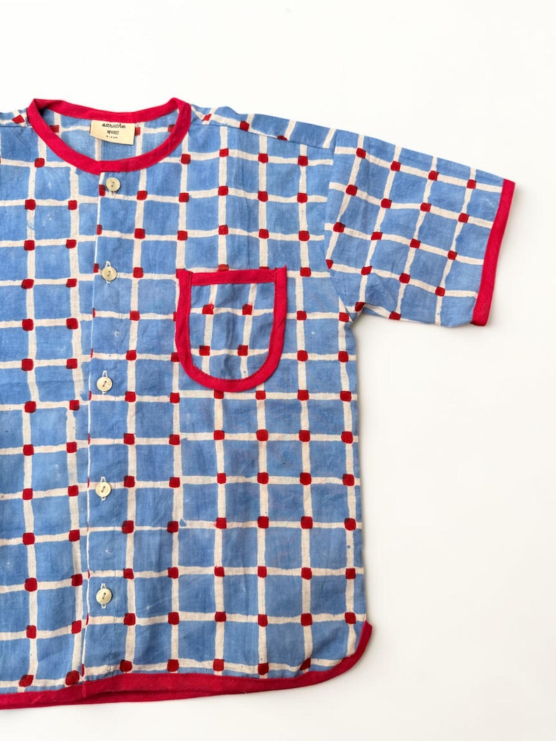 May include: A short-sleeved button-down shirt featuring a light blue grid pattern on a cream background with small red squares at each intersection. The shirt has a red collar, red sleeve cuffs, a red hem, and a single chest pocket with red trim.
