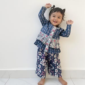 May include: A young child wearing a matching two-piece outfit. The top is a navy blue jacket with a floral pattern and a ruffled hem. The pants are wide-legged and feature a similar floral design. The child is smiling and has their arms raised.