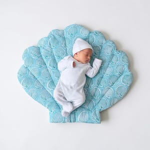 Peut inclure: Un nouveau-né portant un body blanc à manches longues et un bonnet blanc assorti, allongé sur un grand tapis rembourré en forme de coquillage. Le tapis présente un motif répétitif de coquillages bleu sarcelle sur un fond en tissu bleu clair.