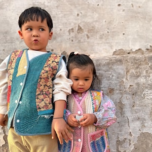 May include: Two children in patchwork vests. The boy wears a teal vest over a white top and beige trousers. The girl wears a pink vest over a floral dress. The vests feature a quilted design with diverse fabric patterns.