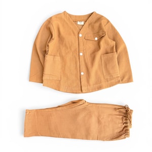 May include: A two-piece set of children's clothing in a warm tan color. The top is a long-sleeved jacket with a V-neck, two front pockets, and white buttons. The matching pants have an elastic waistband and drawstring. The fabric has a waffle-like texture.