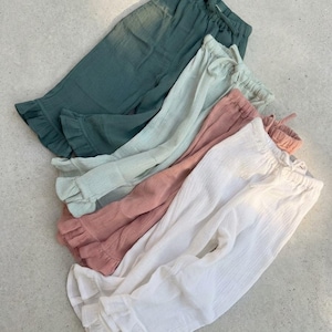 May include: A collection of five pairs of lightweight, ruffled-hem pants in various colors. The colors include teal, mint green, dusty rose, and white. Each pair has a drawstring waistband.