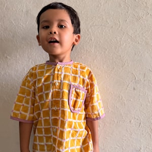 May include: A short-sleeved button-down shirt featuring a yellow and white grid pattern with light purple trim on the collar, sleeve cuffs, and chest pocket. The shirt has a mandarin collar and white buttons running down the center front.