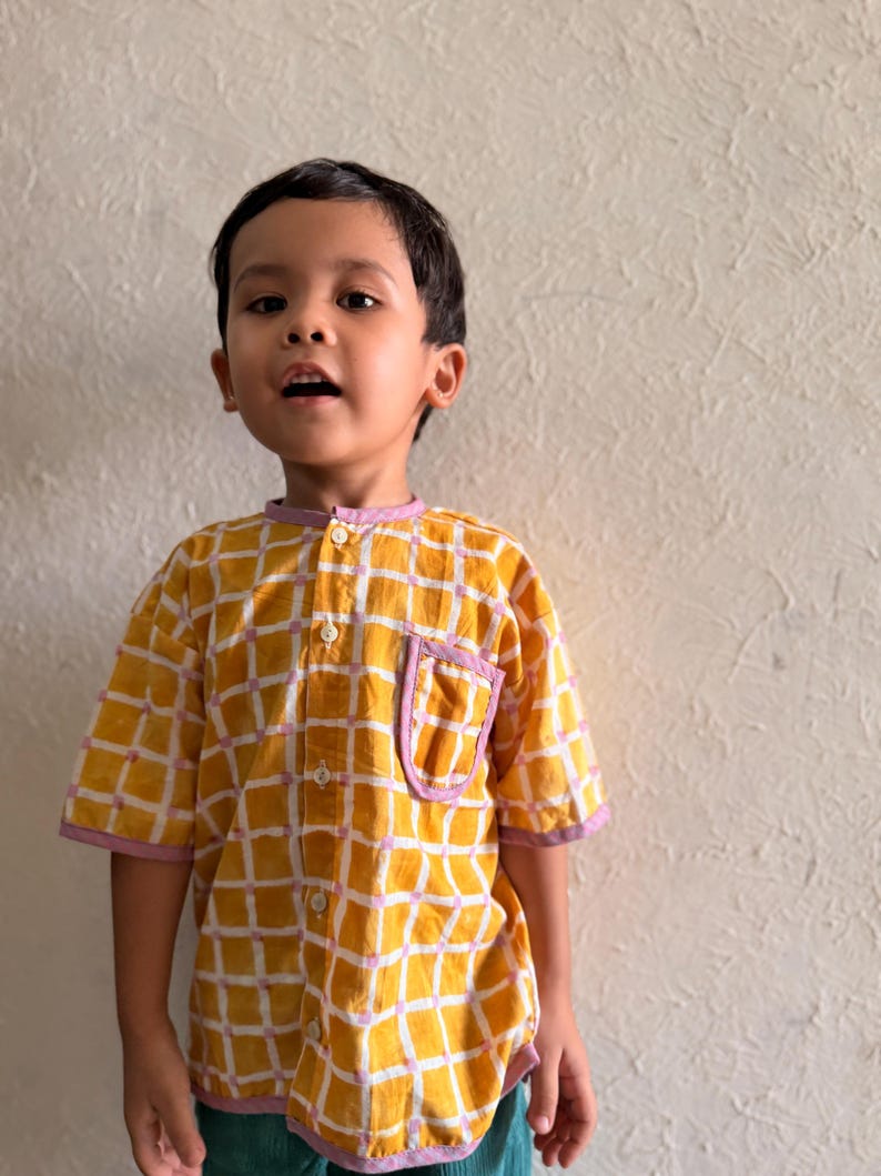 May include: A short-sleeved button-down shirt featuring a yellow and white grid pattern with light purple trim on the collar, sleeve cuffs, and chest pocket. The shirt has a mandarin collar and white buttons running down the center front.