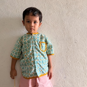 May include: A short-sleeved button-down shirt featuring a light green and white grid pattern with mustard yellow accents on the collar, sleeve cuffs, pocket, and hem, paired with solid light pink shorts that reach above the knee.