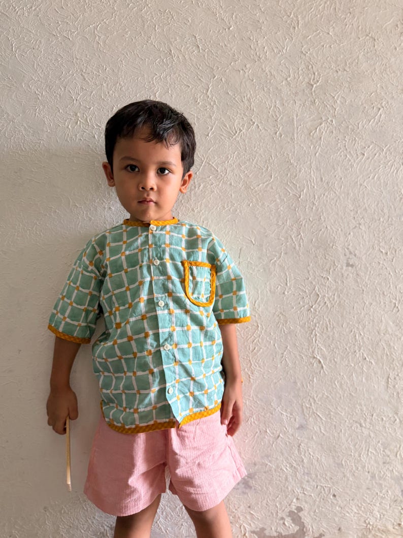 May include: A short-sleeved button-down shirt featuring a light green and white grid pattern with mustard yellow accents on the collar, sleeve cuffs, pocket, and hem, paired with solid light pink shorts that reach above the knee.