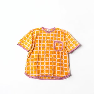 May include: A short-sleeved button-down shirt featuring a vibrant orange and white grid pattern with light purple trim along the collar, sleeve cuffs, hem, and single chest pocket. The shirt includes a row of small, light-colored buttons down the front.