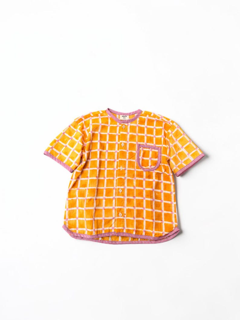 May include: A short-sleeved button-down shirt featuring a vibrant orange and white grid pattern with light purple trim along the collar, sleeve cuffs, hem, and single chest pocket. The shirt includes a row of small, light-colored buttons down the front.