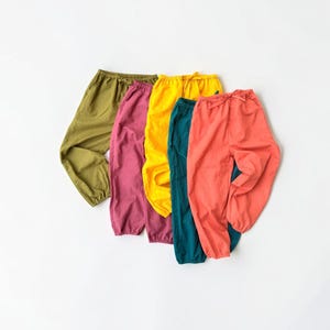 May include: A collection of five pairs of loose-fitting trousers in olive green, magenta, bright yellow, teal, and coral. Each pair features an elasticated waistband with a drawstring tie and elastic cuffs at the ankles, creating a gathered look.