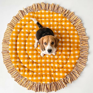 Pode incluir: Um tapete redondo e acolchoado para animais de estimação com um padrão xadrez vichy em amarelo mostarda e branco. O tapete tem uma borda com babados e um design listrado em marrom, branco e amarelo mostarda. Um beagle está sentado no tapete.