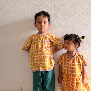 May include: A yellow and white grid-patterned button-down shirt with a lavender trim and a chest pocket, paired with solid dark green trousers, alongside a matching yellow and white grid-patterned dress with a lavender trim and button front.