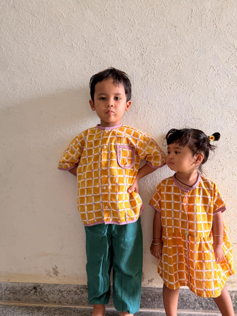 May include: A yellow and white grid-patterned button-down shirt with a lavender trim and a chest pocket, paired with solid dark green trousers, alongside a matching yellow and white grid-patterned dress with a lavender trim and button front.