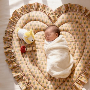 Może przedstawiać: Śpiące dziecko owinięte w biały kocyk na sercowatej, musztardowo-żółtej macie dla dziecka z kwiatowym wzorem. Biały i żółty pluszowy lew oraz czerwona i drewniana zabawka leżą obok dziecka. Mata ma falbankę.