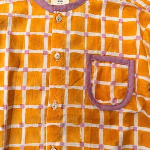 May include: A long-sleeved shirt featuring a yellow and white grid pattern with small pink squares at the intersections. The shirt has a round neckline, a front button placket with four light-colored buttons, and a chest pocket with purple trim.