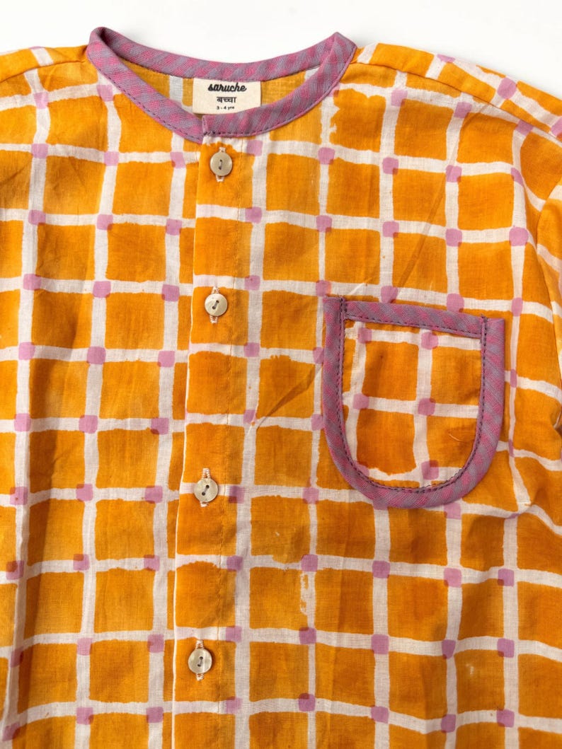 May include: A long-sleeved shirt featuring a yellow and white grid pattern with small pink squares at the intersections. The shirt has a round neckline, a front button placket with four light-colored buttons, and a chest pocket with purple trim.
