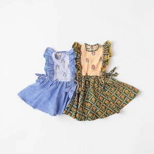 Op de afbeelding: Twee mouwloze jurken met ruches op de schouders en strikbandjes aan de zijkant. De linkerjurk heeft een blauw-wit gestreept lijfje met een bloemenprint en een blauwe rok met vichy-ruitjes. De rechterjurk heeft een geel-wit gestreept lijfje met een bloemenprint en een groene rok met een oranje bloemenpatroon.