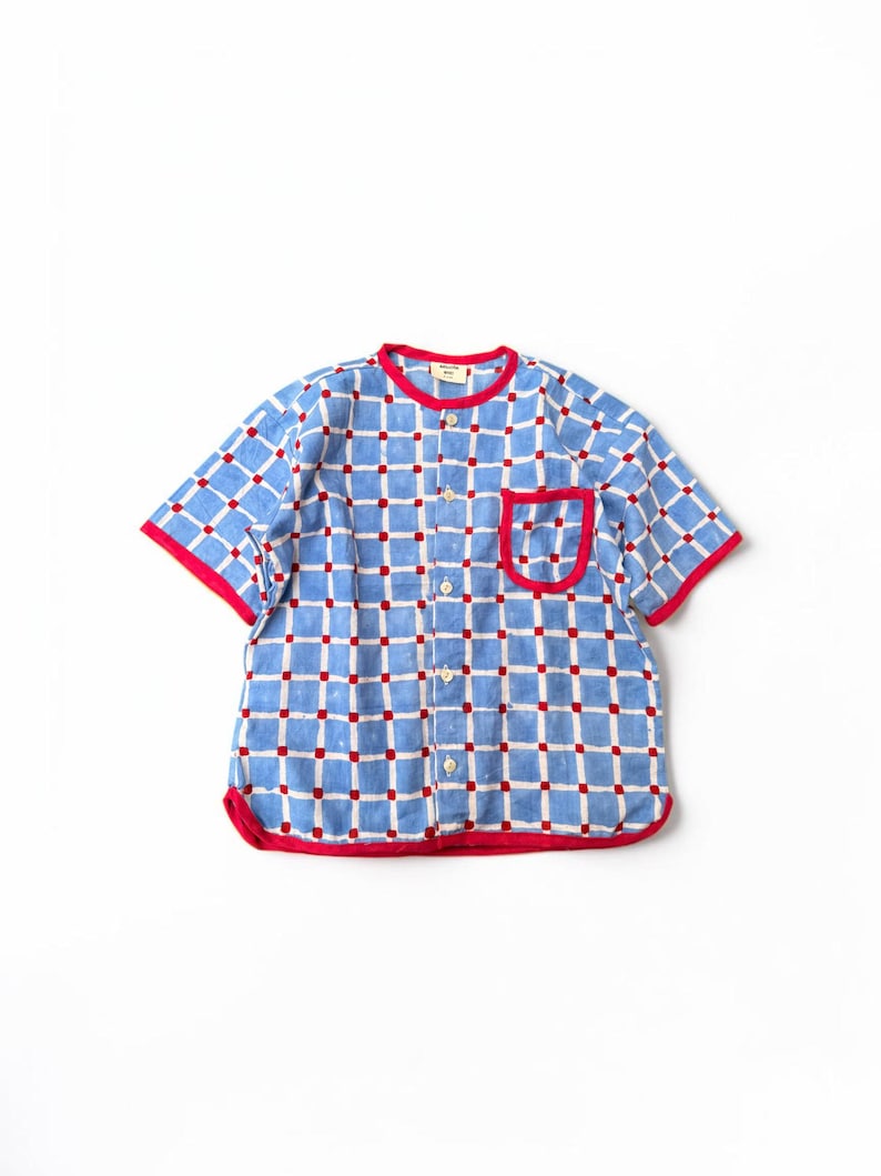 May include: A short-sleeved button-down shirt featuring a blue and white grid pattern with small red squares at each intersection. The shirt has a round neckline, a single chest pocket, and bright red trim along the collar, pocket, and hem.