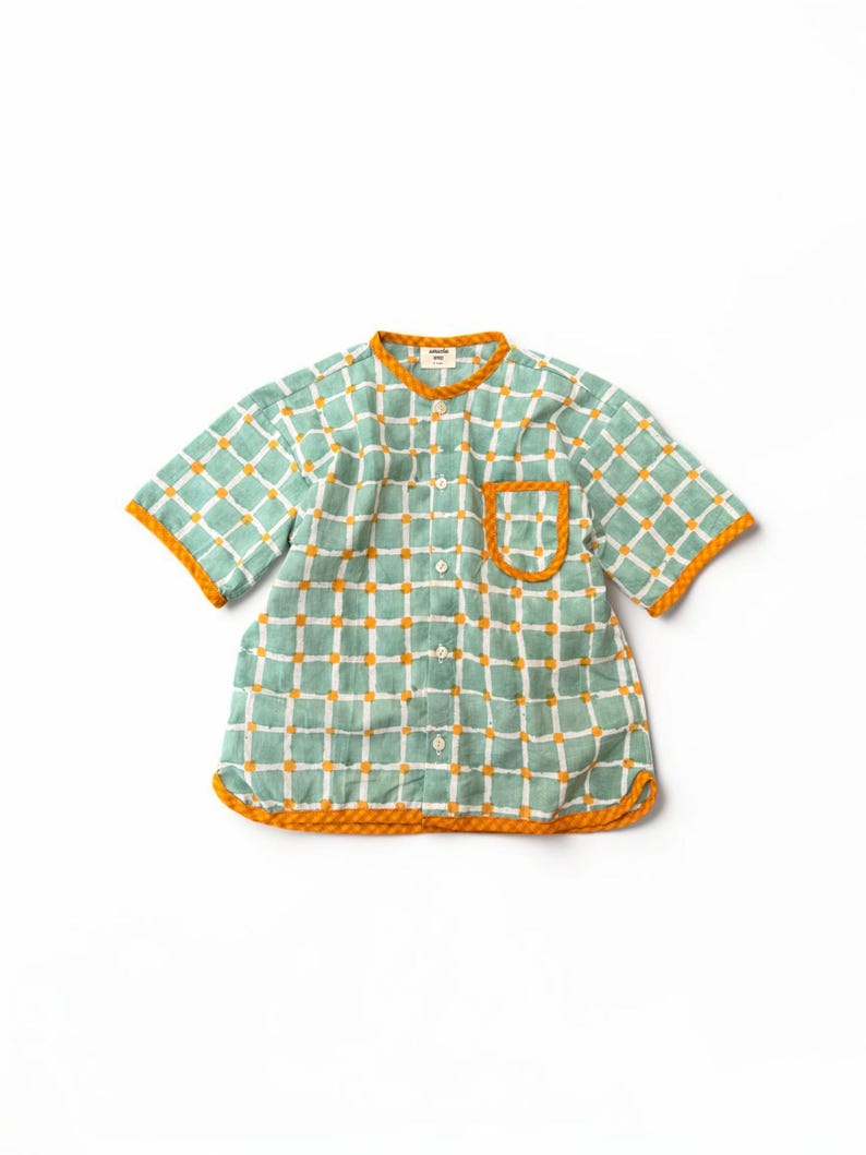 May include: A short-sleeved button-down shirt featuring a light green and white grid pattern with small orange squares at the intersections. The shirt has an orange trim along the collar, sleeve cuffs, hem, and the single chest pocket.