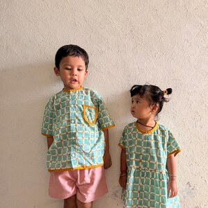 May include: A short-sleeved button-down shirt with a light green and white grid pattern and mustard yellow trim, paired with pink shorts, alongside a short-sleeved dress featuring the same green and white grid pattern and mustard yellow trim.