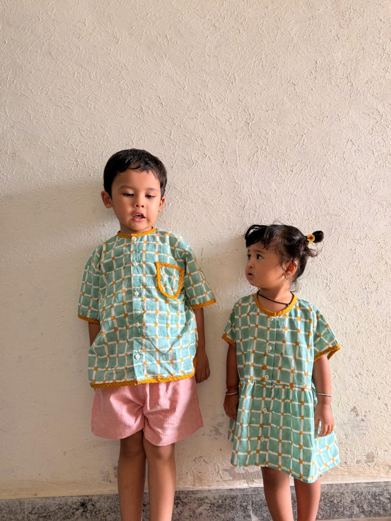 May include: A short-sleeved button-down shirt with a light green and white grid pattern and mustard yellow trim, paired with pink shorts, alongside a short-sleeved dress featuring the same green and white grid pattern and mustard yellow trim.